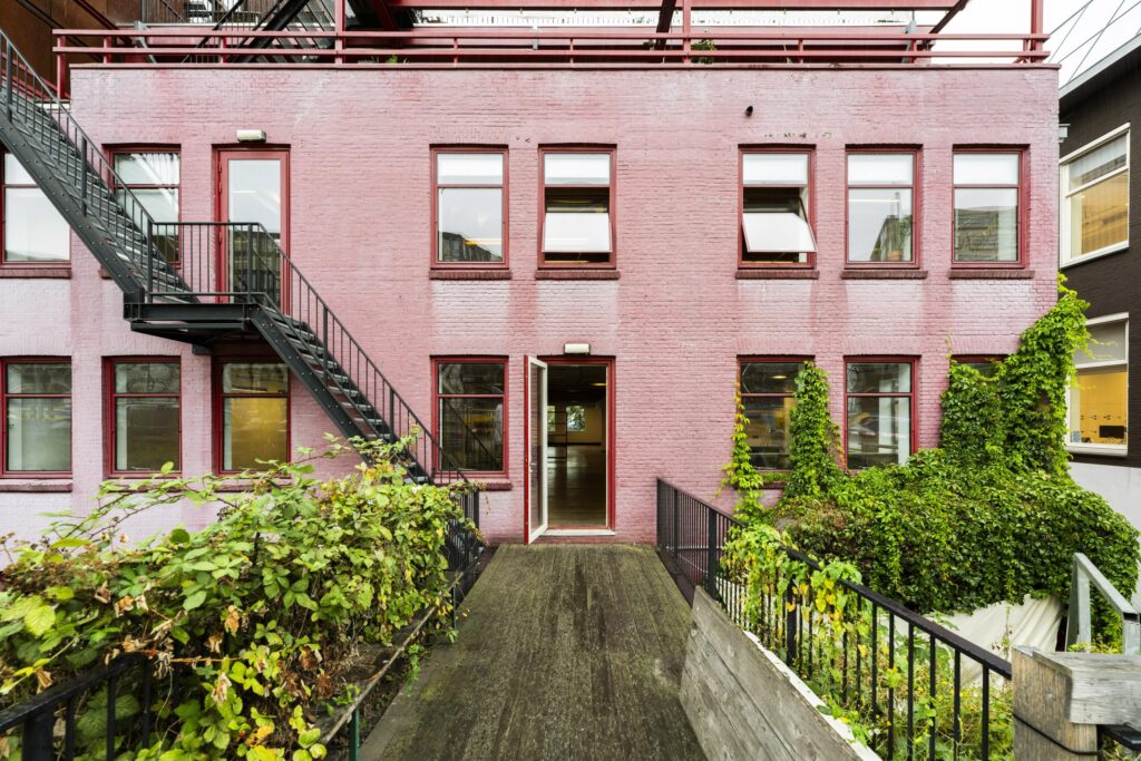 Roze bakstenen gevel van een gebouw aan de De Ruijterkade met openstaande deur, metalen trap en begroeide planten.